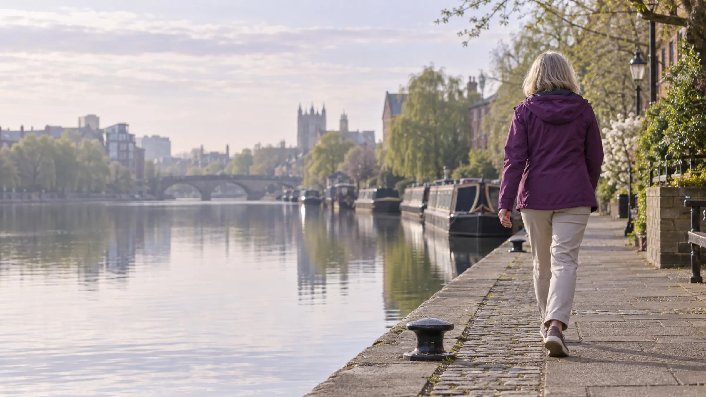 A woman walking briskly along a canal towpath in soft morning light, seen from behind