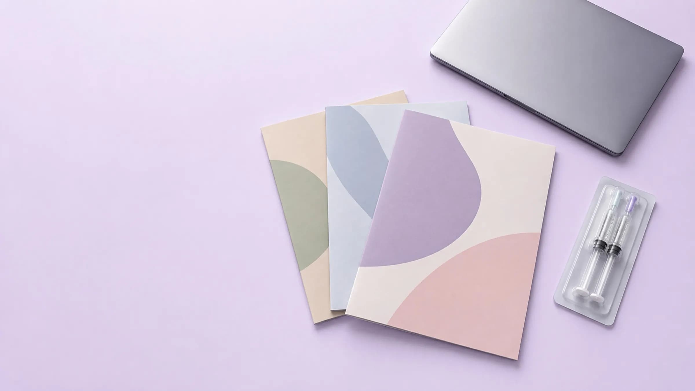 Three brochures fanned on a lilac surface beside a laptop and a syringe in its blister